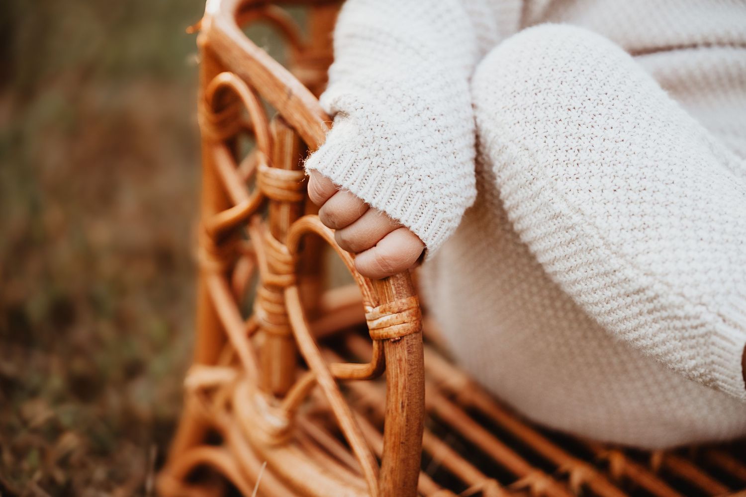 Close-up of a baby hand resting on a wicker basket wrapped in a white knit blanket outdoors.