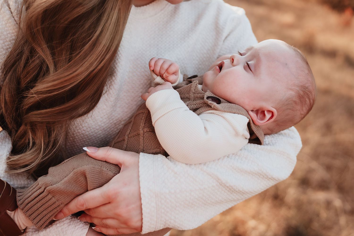Mother in cream sweater cradling her sleeping newborn baby outdoors in warm autumn light.