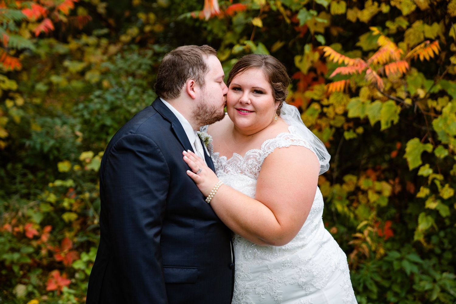 Groom kisses bride on cheek as she smiles before vibrant autumn foliage in wedding portrait.