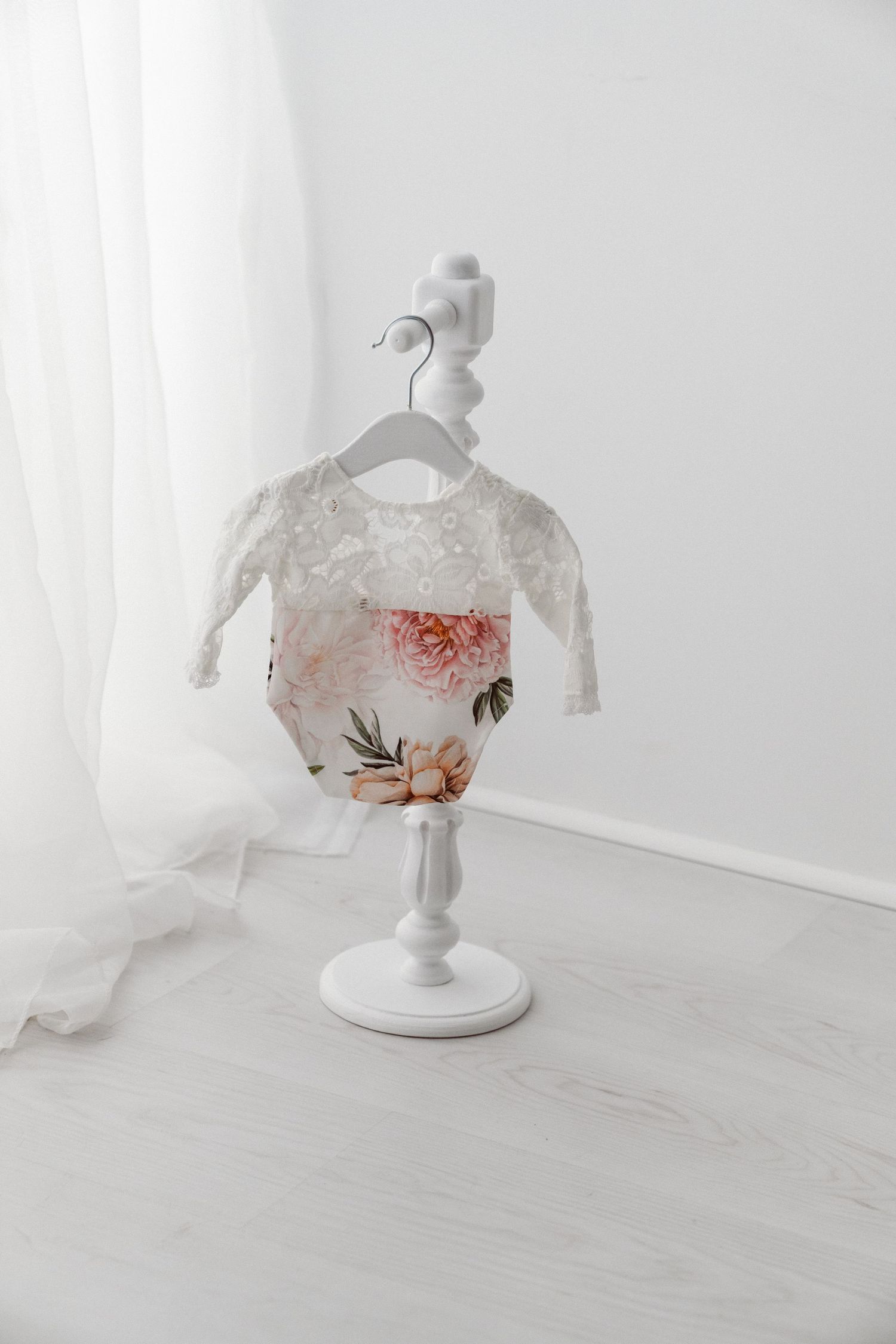 Floral print baby romper with white lace sleeves displayed on a white decorative mini stand in a bright white room.