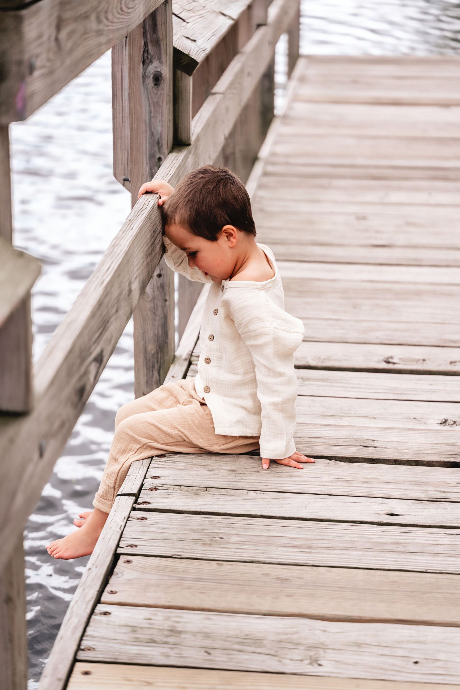 Young barefoot boy in linen outfit sitting on wooden dock railing by water.