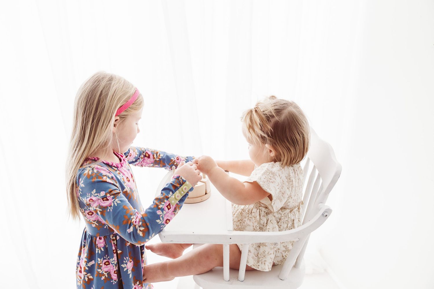 Two young sisters playing together at a white children's table in a bright, airy room.