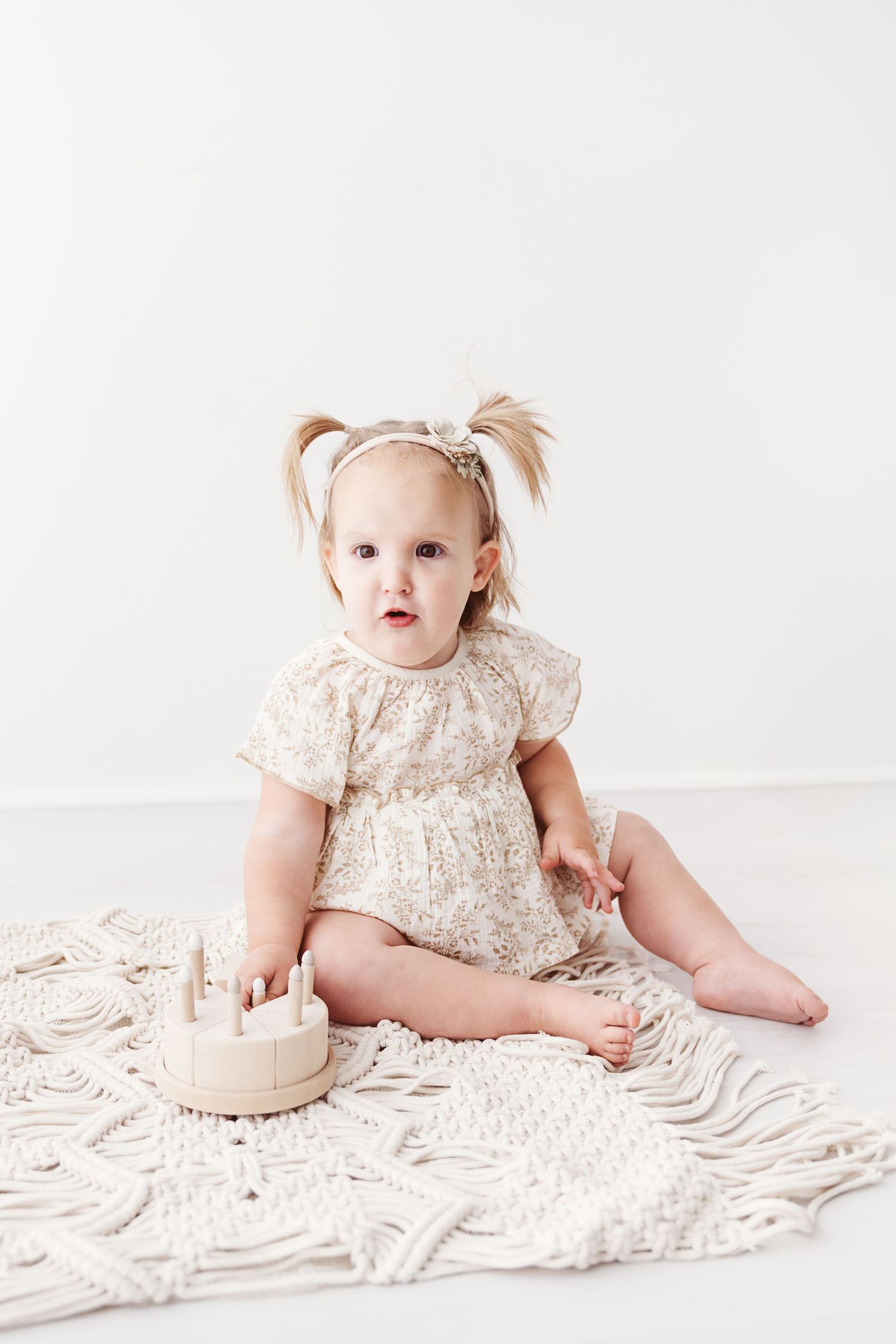 An adorable toddler girl in a floral romper sits on a macrame blanket with a wooden toy cake.
