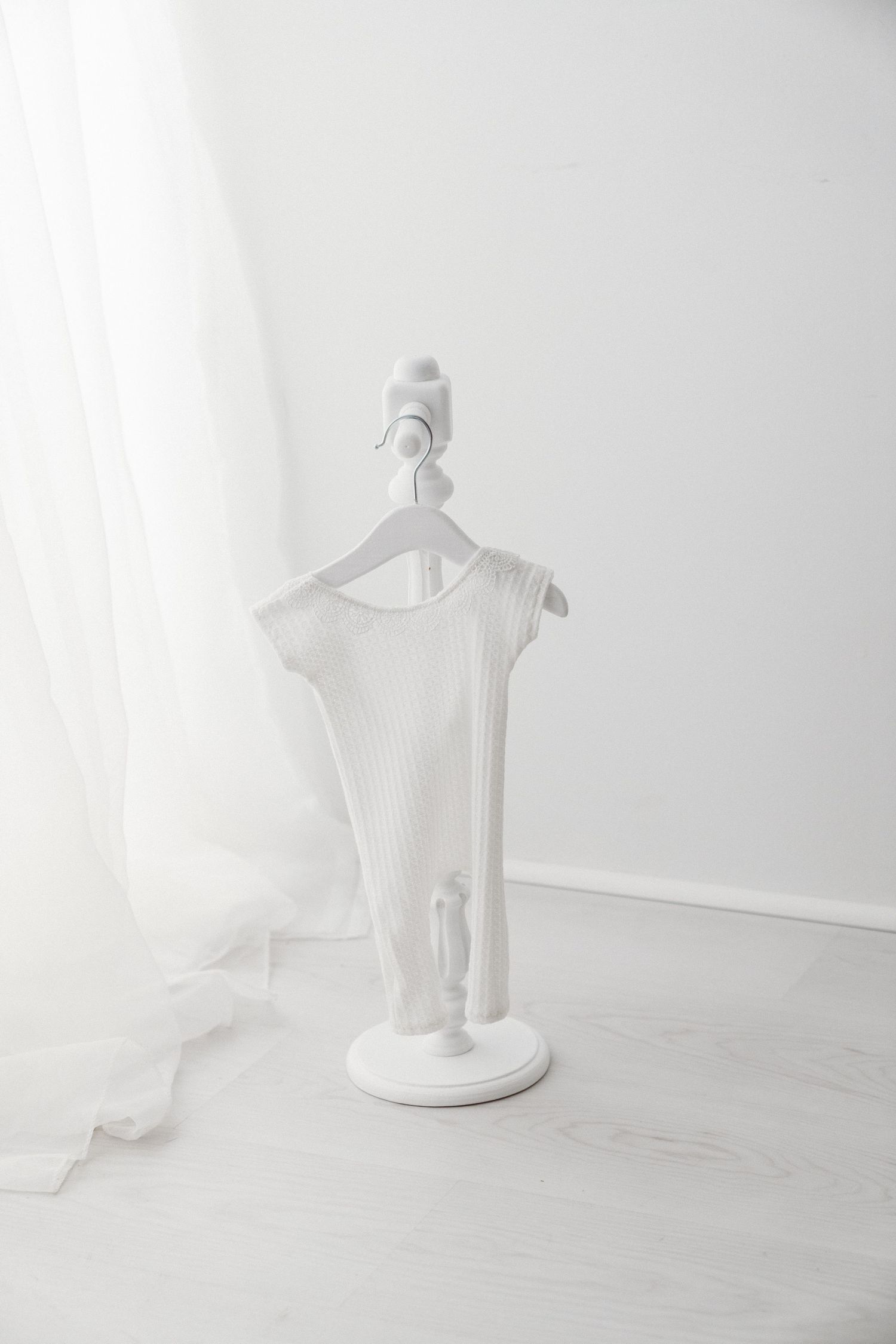 A white ribbed newborn onesie displayed on a miniature white dress form stand by sheer curtains.