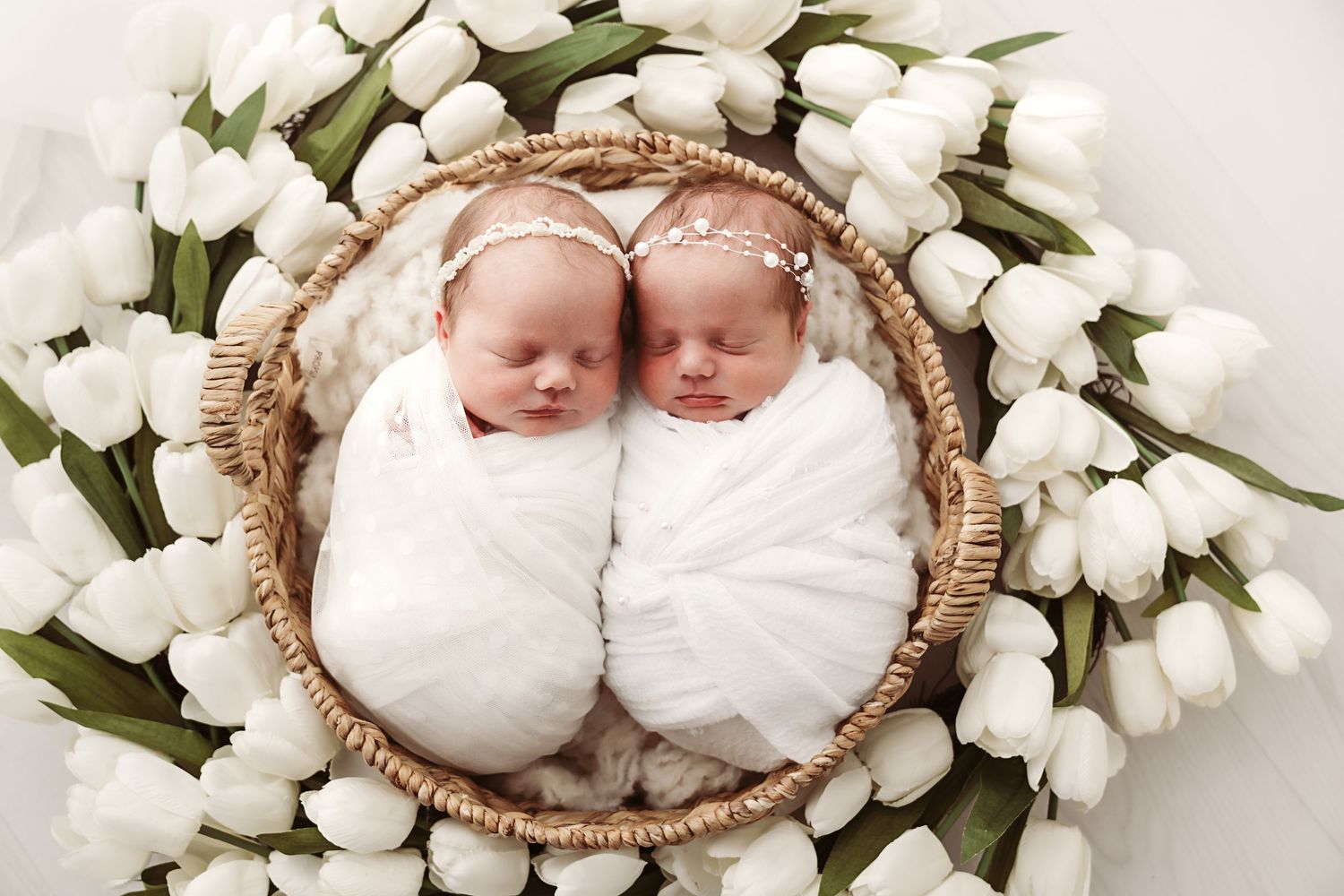 Twin newborns swaddled in white wraps sleeping in a wicker basket surrounded by white tulips.