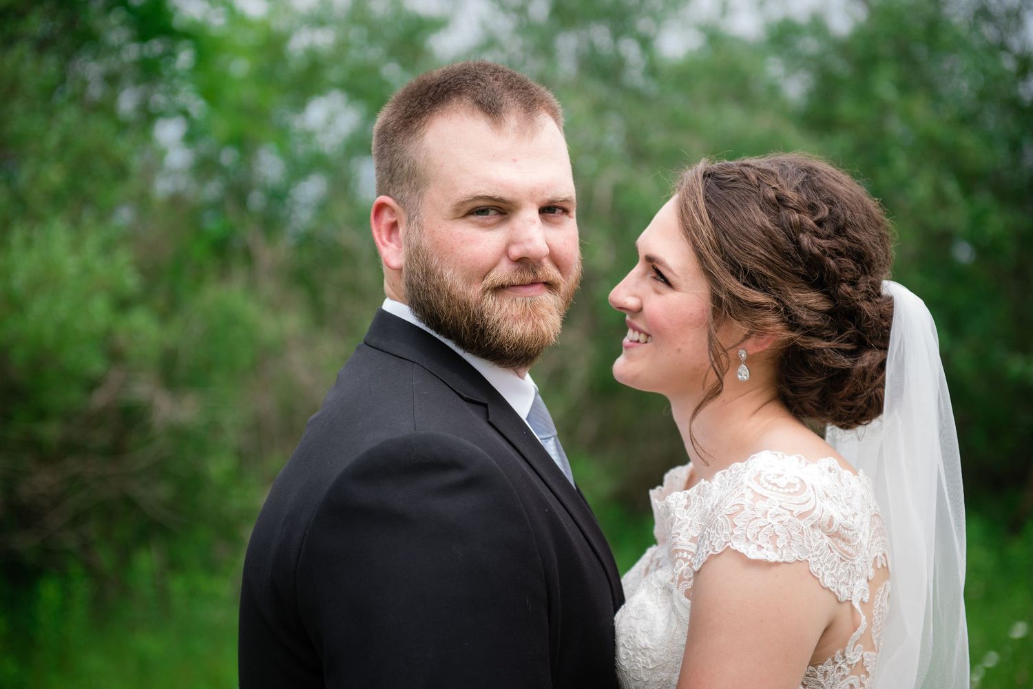 Smiling wedding couple outdoors, groom with beard in dark suit, bride in lace cap-sleeve gown with veil.