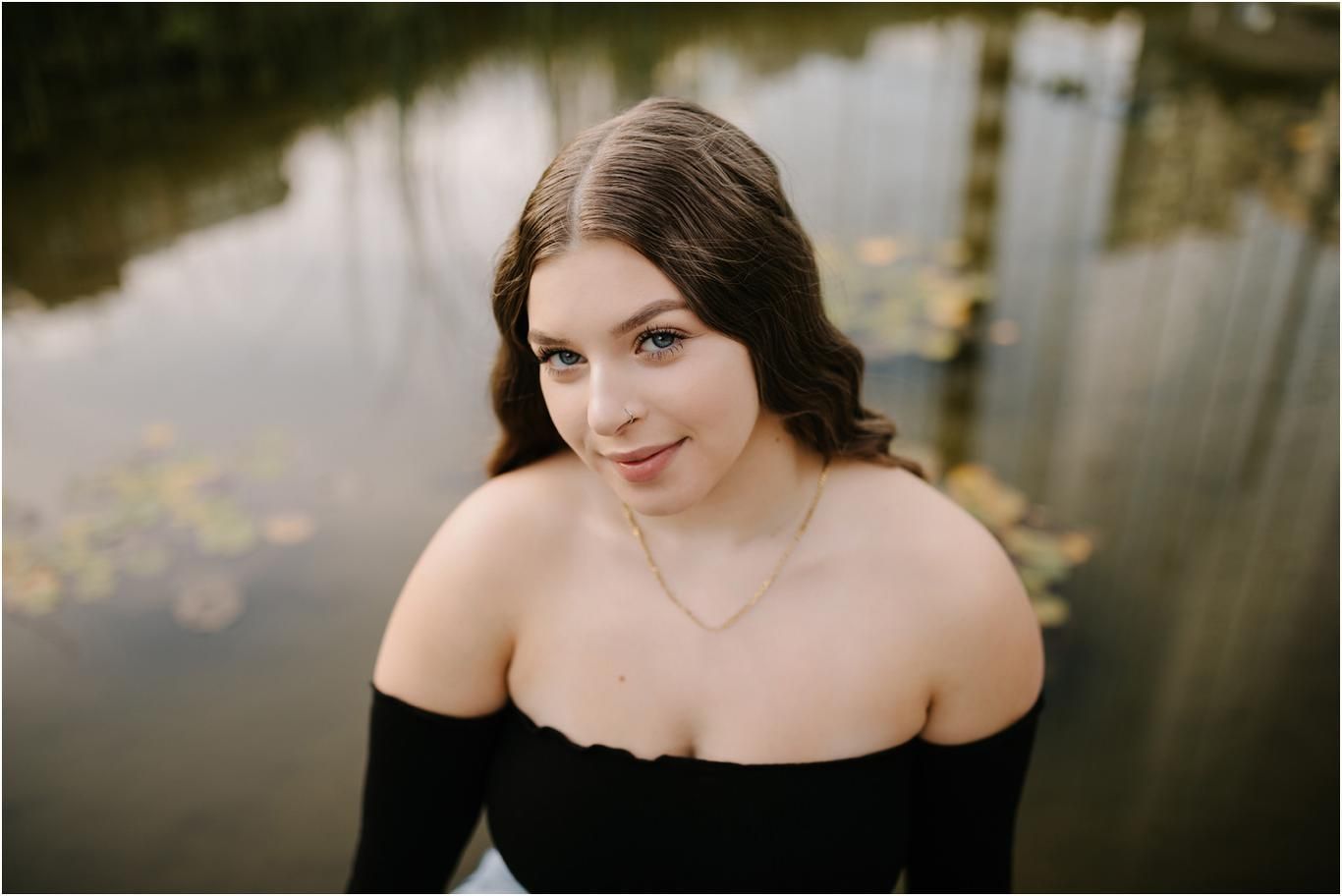 Young woman in black off-shoulder top poses near a reflective pond with lily pads during a senior portrait session.