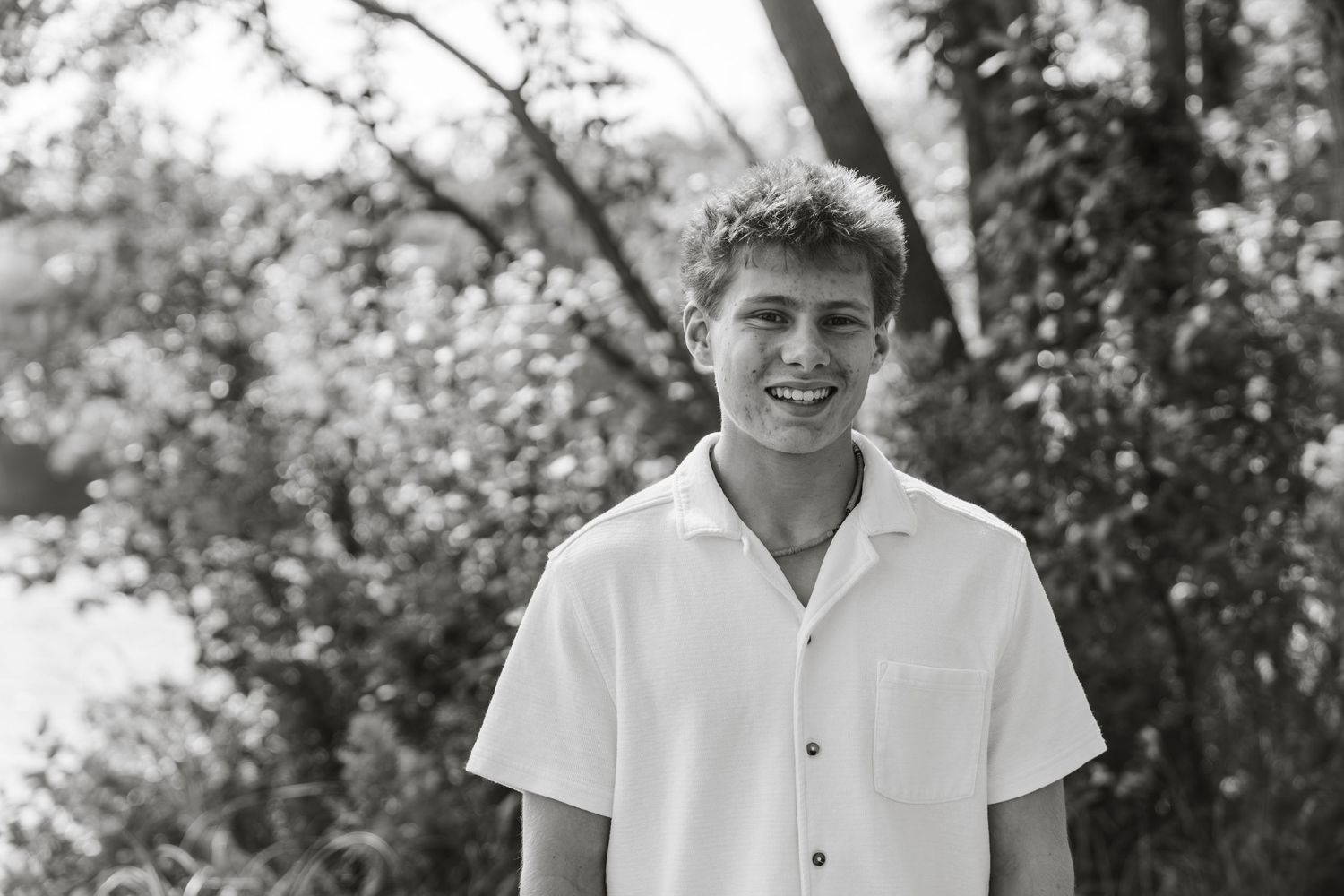 Smiling young man in white button-up shirt outdoors with trees in black and white photo.