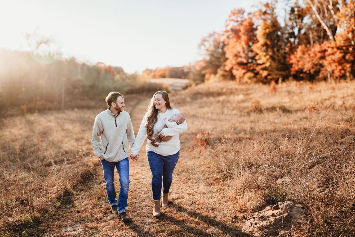 Couple walking hand-in-hand through a golden autumn field, mother carrying their newborn baby.