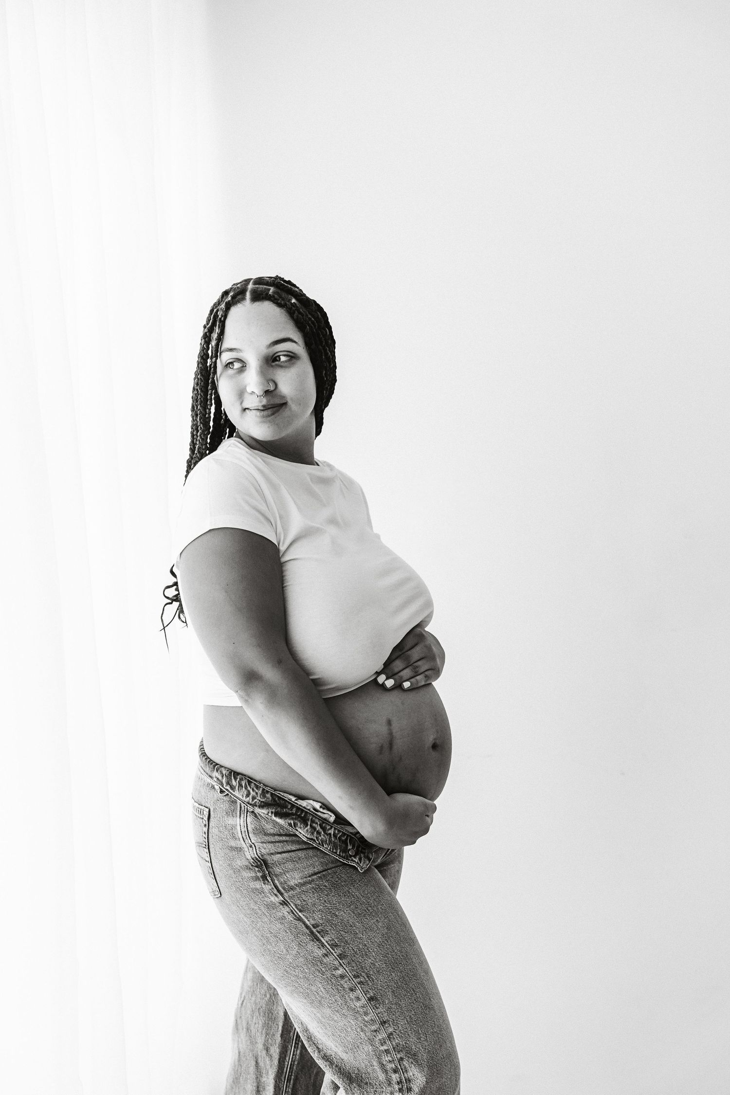 Black and white portrait of pregnant woman in white tee and jeans holding baby bump near curtain.