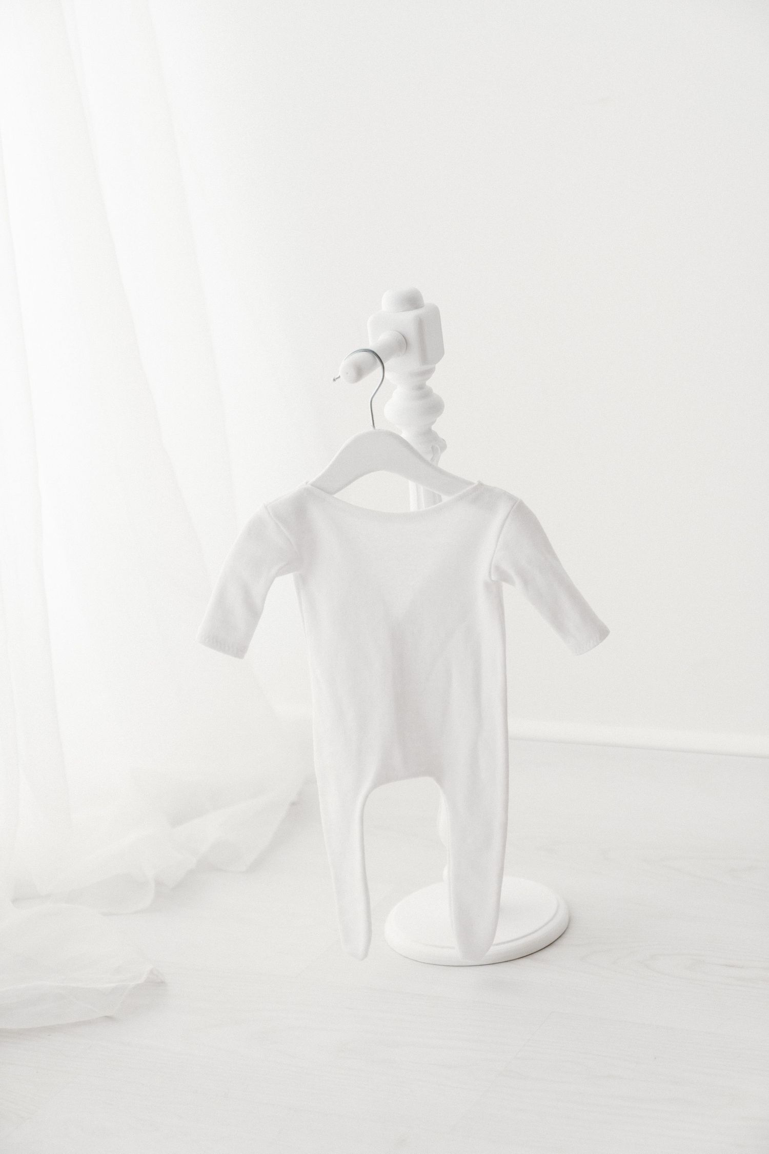White long-sleeve footed baby romper hanging on a white decorative stand in a bright white studio.