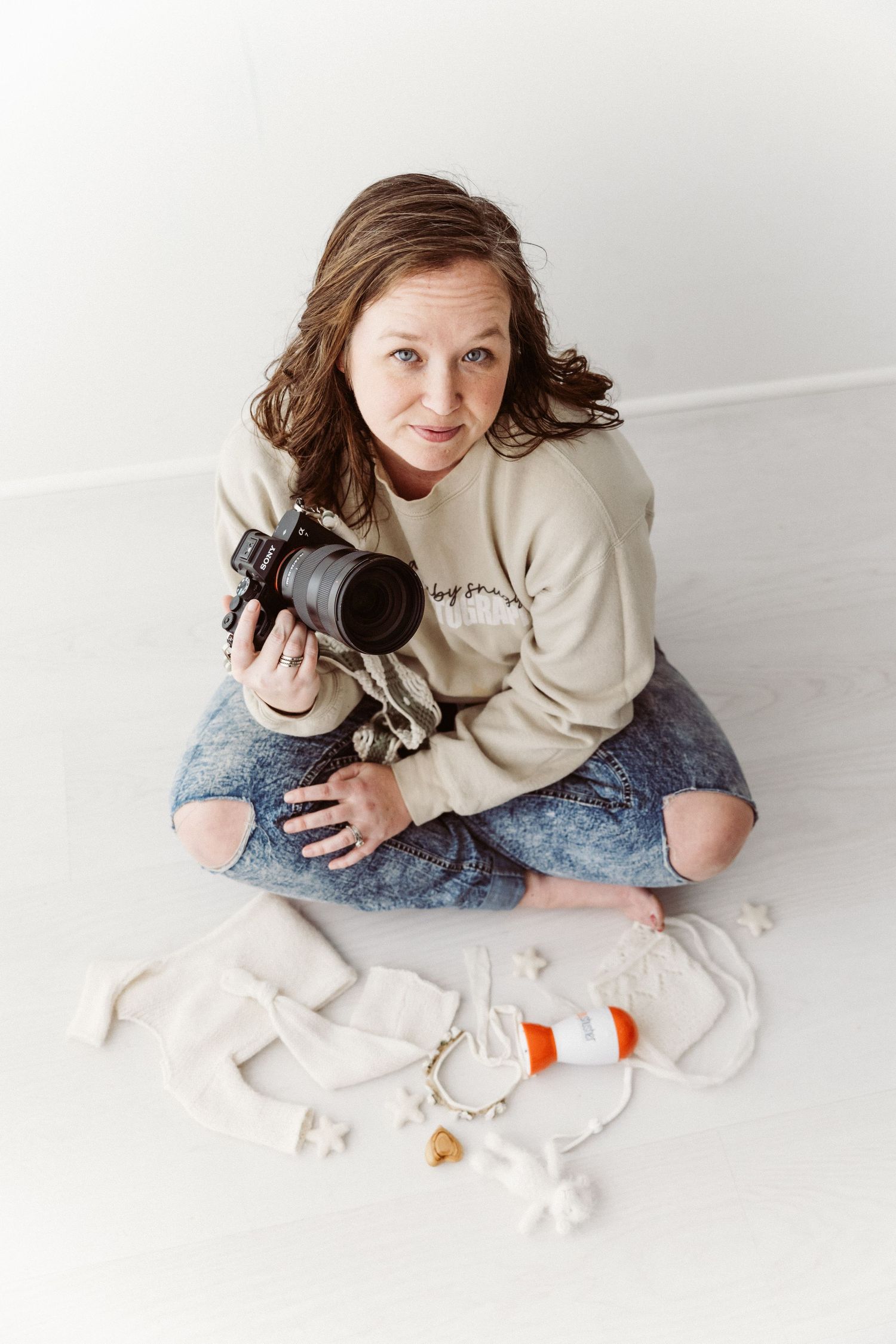 Female photographer sitting cross-legged on white floor holding DSLR camera, with newborn photography props nearby.