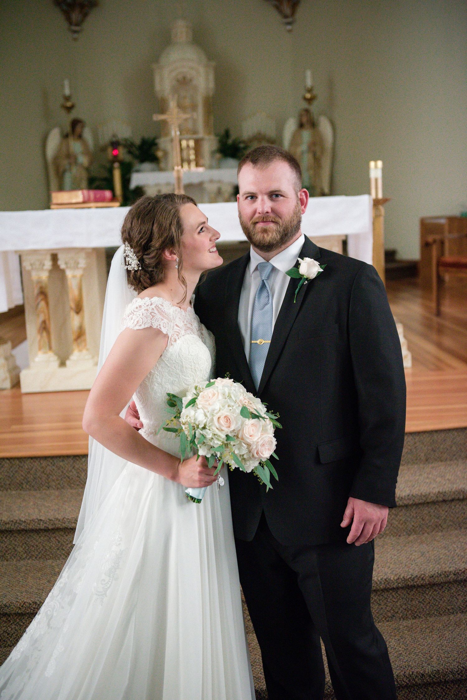 Wedding couple posing at church altar, bride in lace gown holding white rose bouquet, groom in black suit.