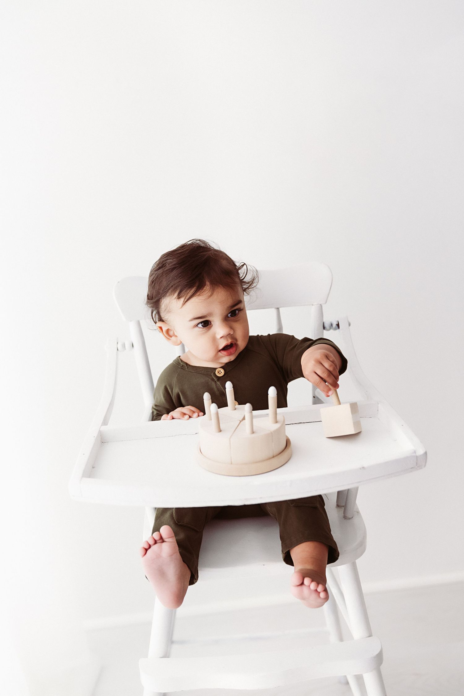 Baby in olive green outfit sitting in white high chair with wooden toy birthday cake.