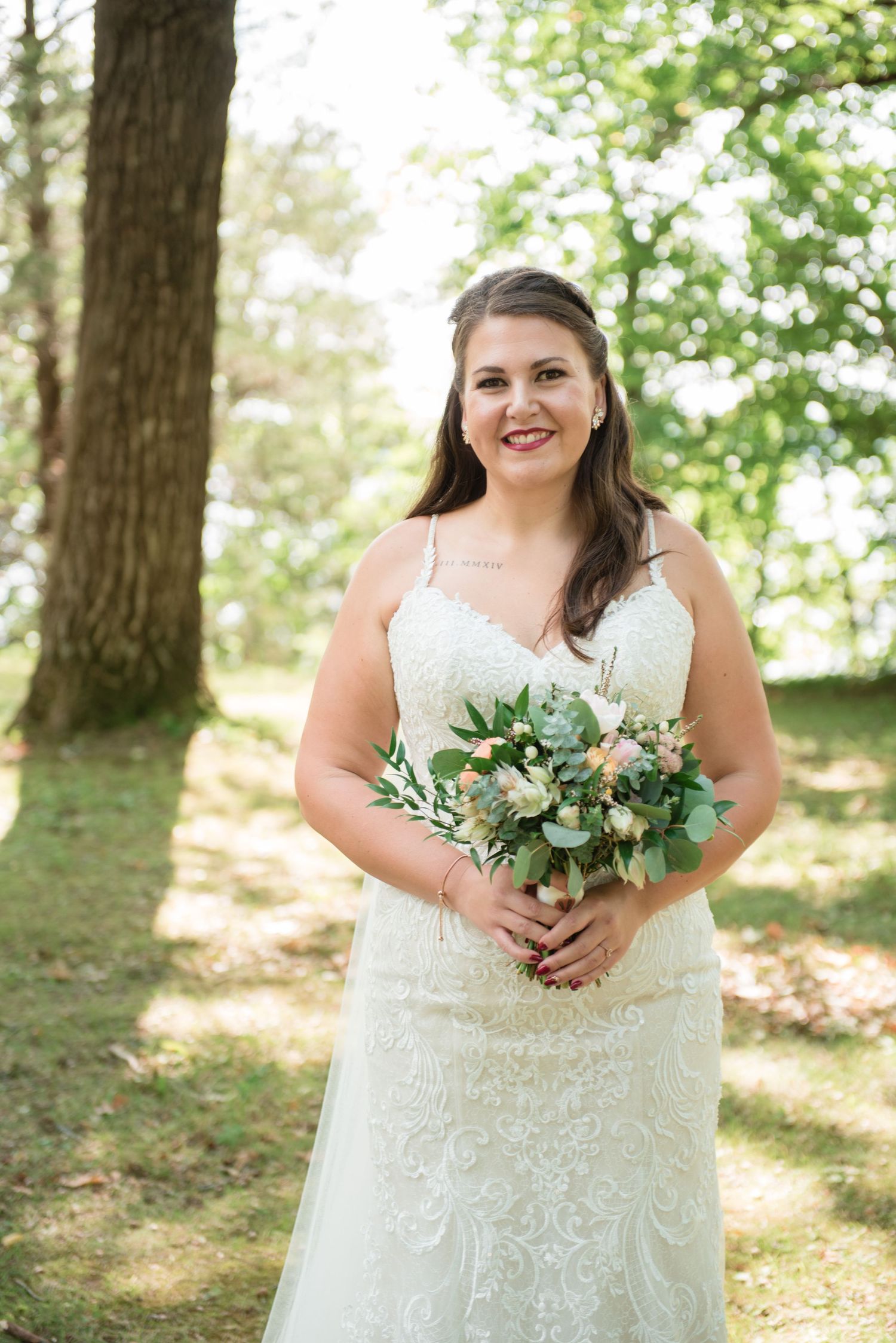 Smiling bride in lace wedding gown holding bouquet of greenery and pink flowers outdoors.