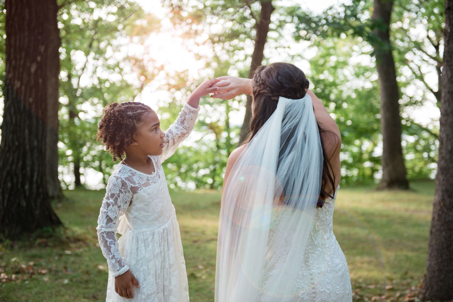 A young flower girl in lace lifts a bride's veil in a sunlit forest during a wedding celebration.
