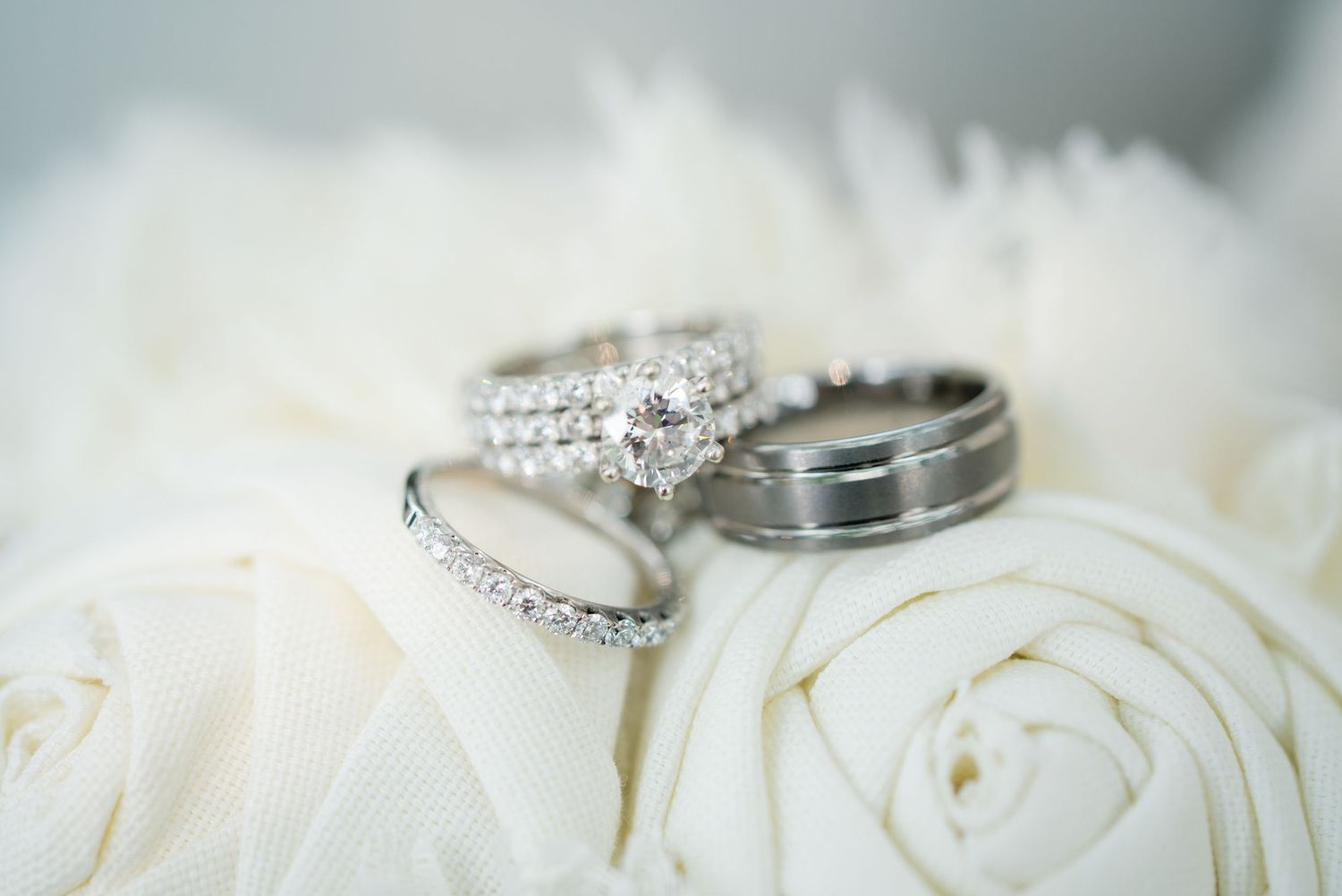 A diamond engagement ring, wedding band, and groom's ring rest elegantly on white rose petals.