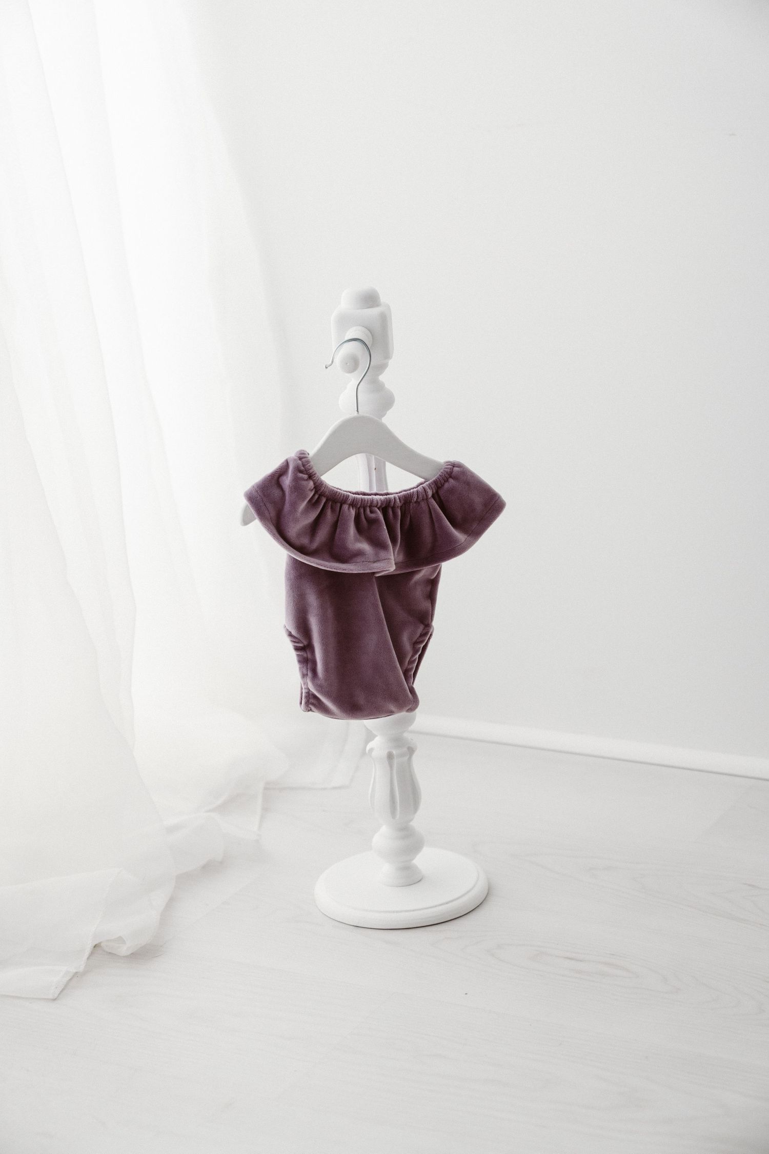 Mauve velvet off-shoulder baby romper with ruffle neckline on a white decorative mini stand.
