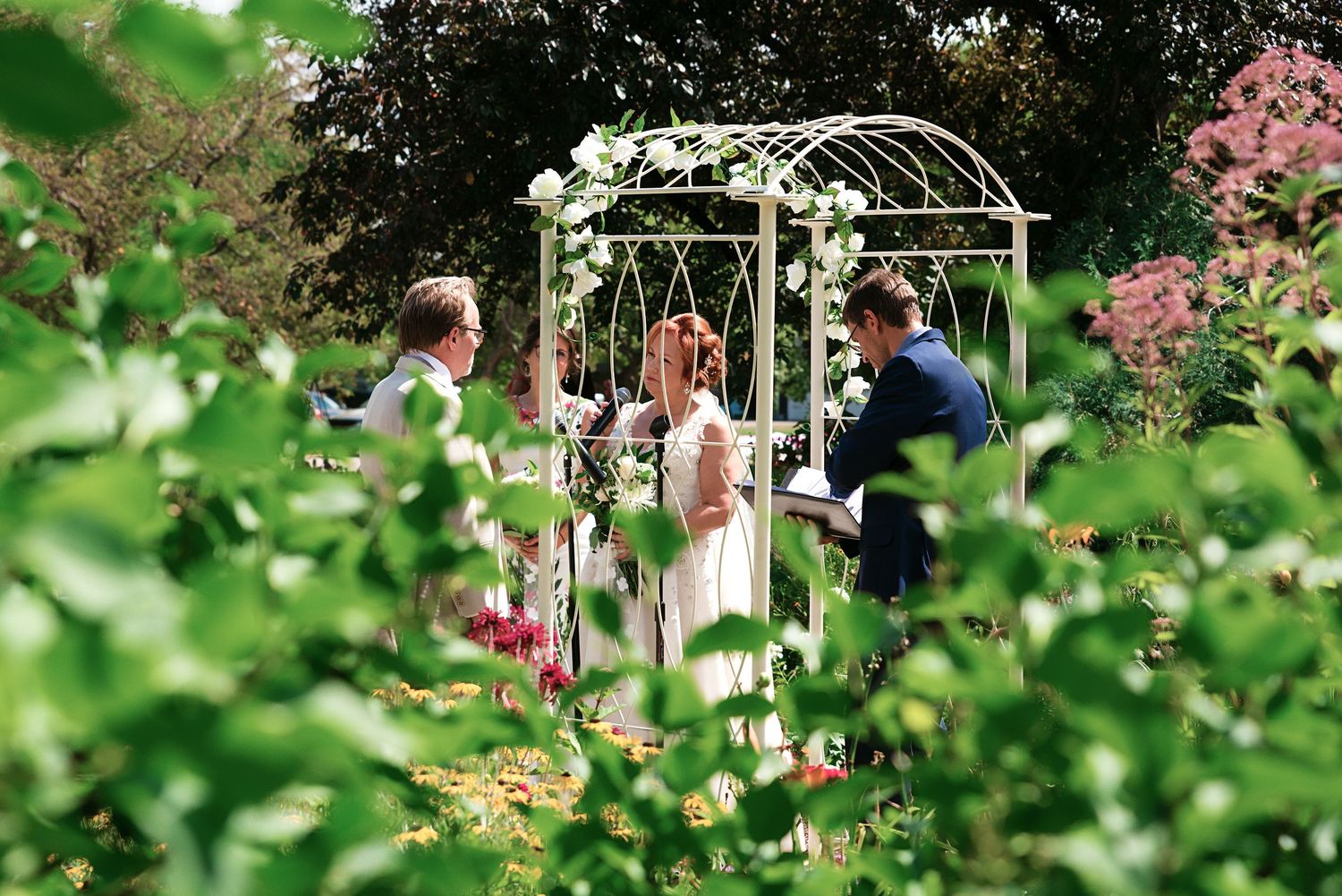 Wedding ceremony in a romantic garden arbor with white roses, officiant and couple surrounded by blooms.