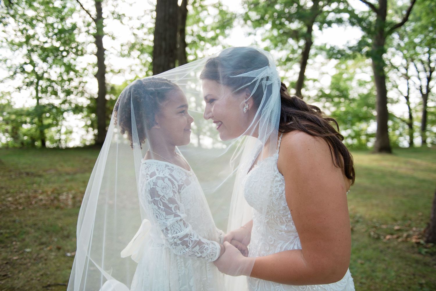 A bride and young flower girl share a tender moment together under a flowing white veil outdoors.