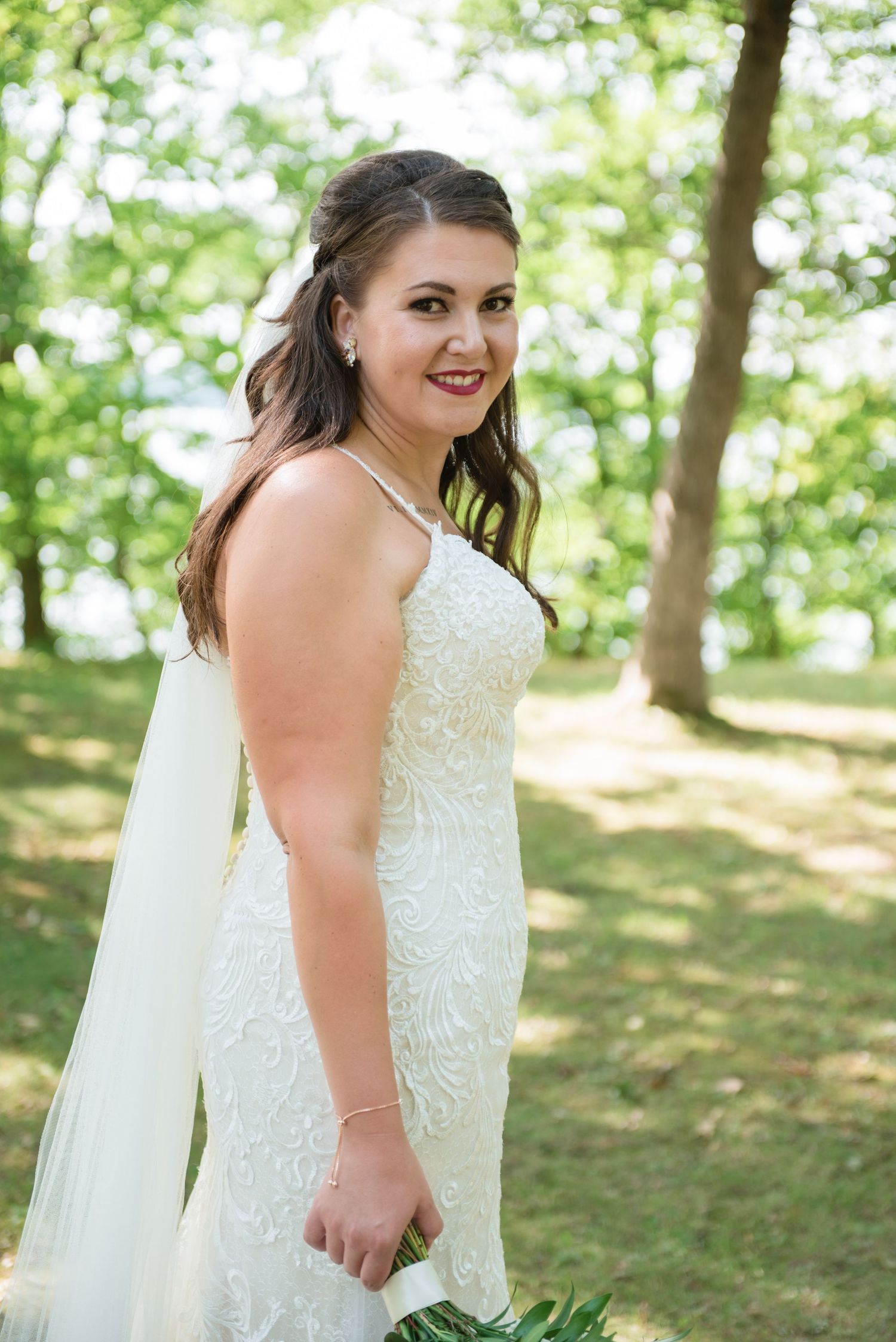 Bride in fitted lace wedding dress with veil smiling over shoulder in sunlit park.