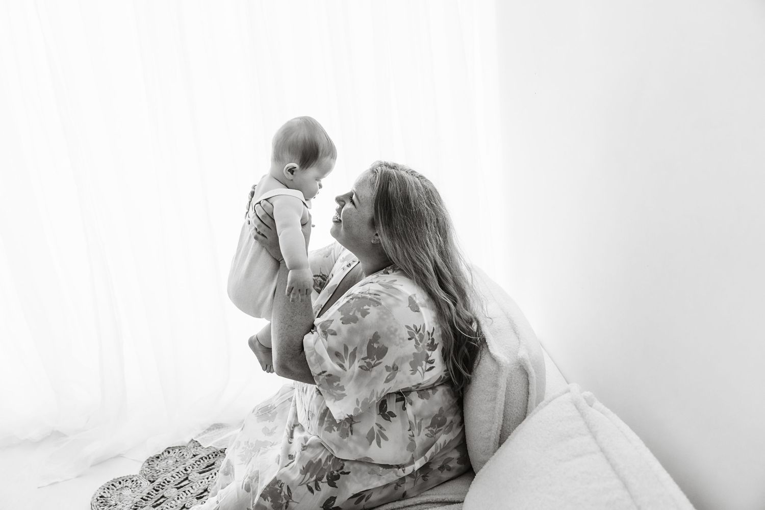 Black and white photo of a mother sitting on a bed holding her baby up near a bright window.