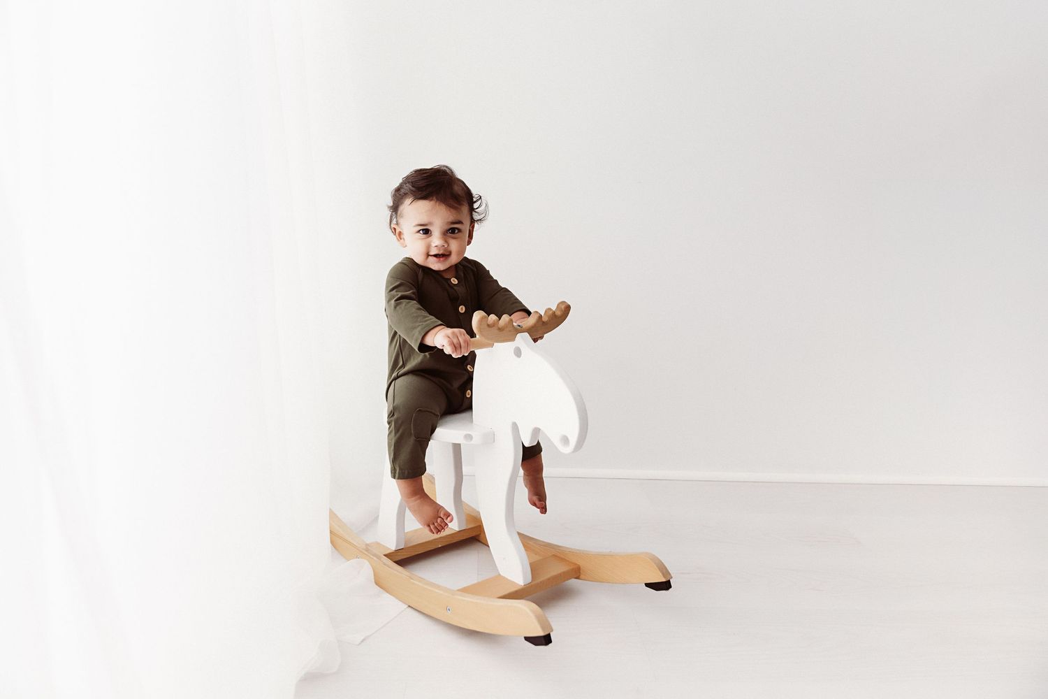 Smiling baby in olive romper riding a white wooden rocking moose toy.