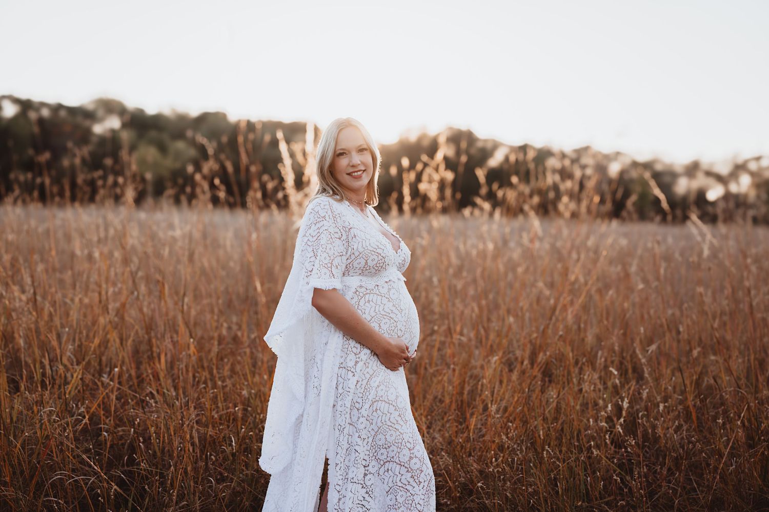 Pregnant woman in a white lace dress cradling her baby bump while standing in a golden field at sunset.