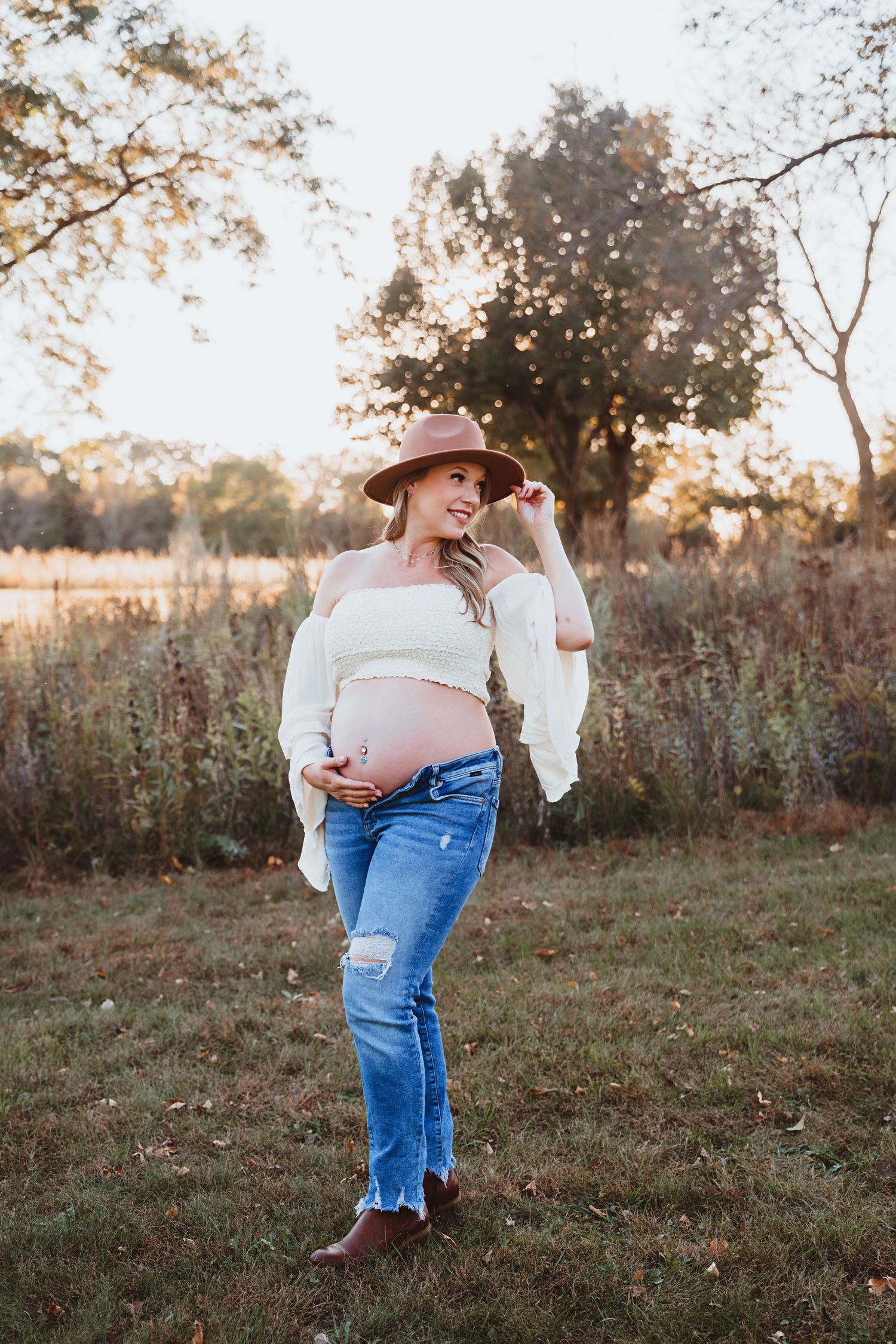 Smiling pregnant woman in a white off-shoulder top, ripped jeans, and brown hat posing outdoors at sunset.