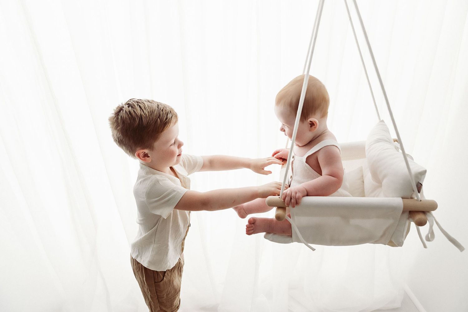 A young boy gently pushes a smiling baby in a white hanging swing indoors.