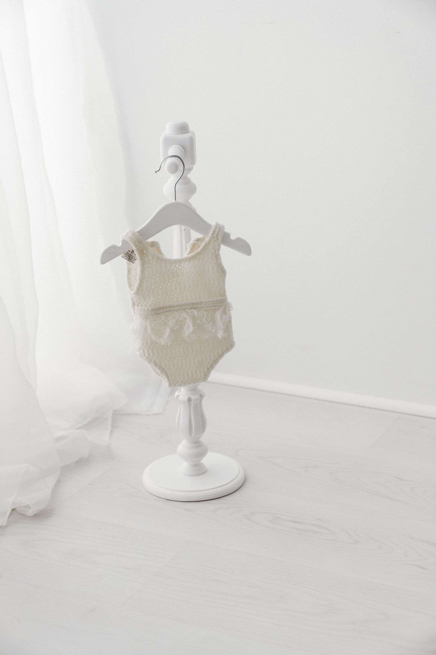 Cream knitted baby romper with fringe detail hanging on a white decorative mini stand in a bright minimalist room.