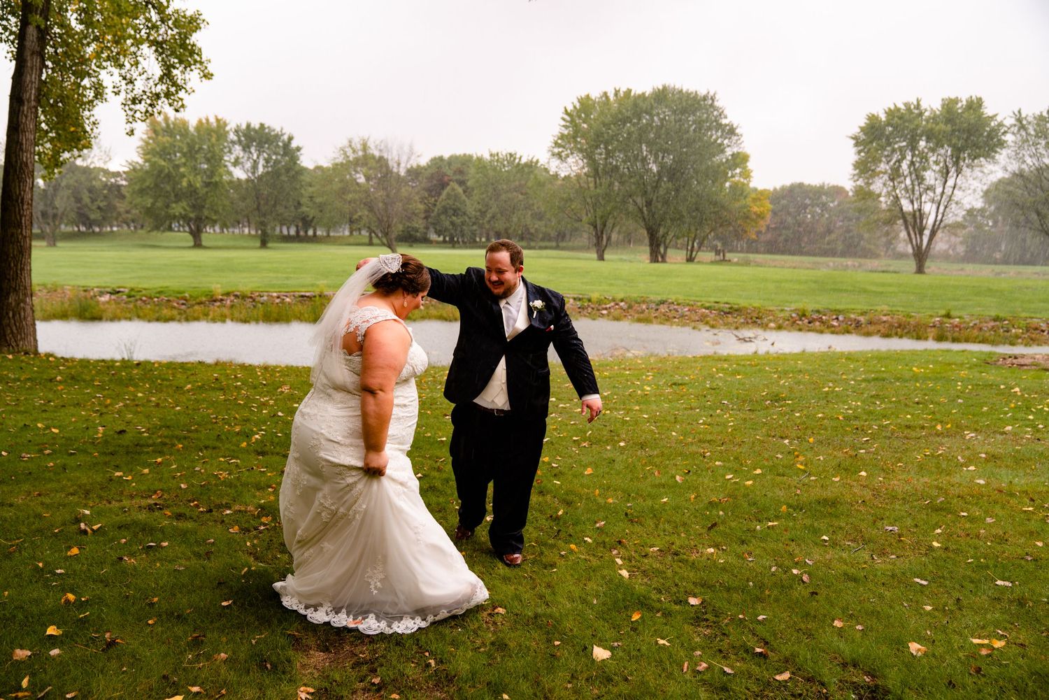 Groom helping bride walk across green lawn on a rainy autumn wedding day near a pond.
