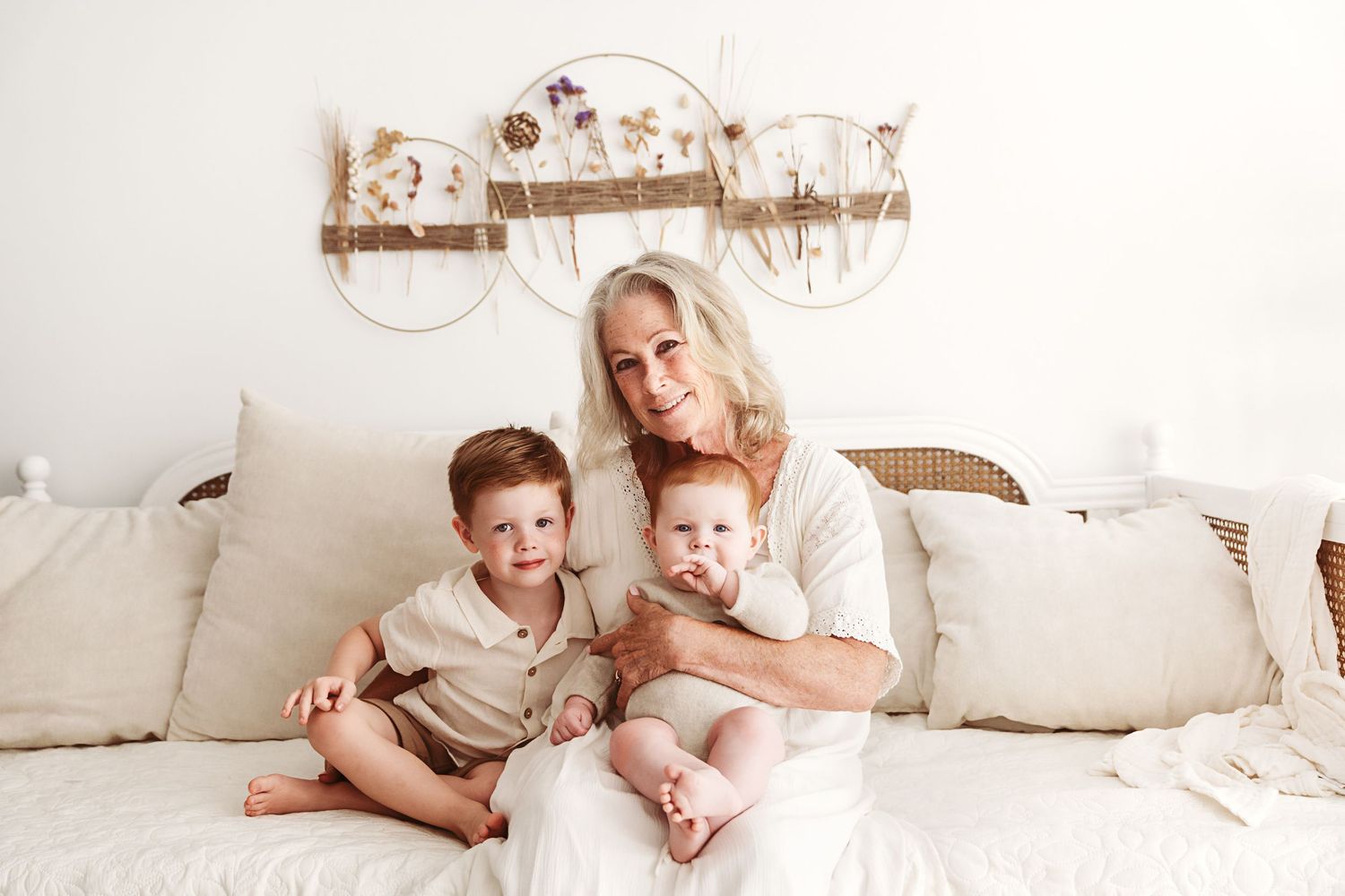 A grandmother poses with her two young grandchildren on a white sofa in a bright boho-styled room.