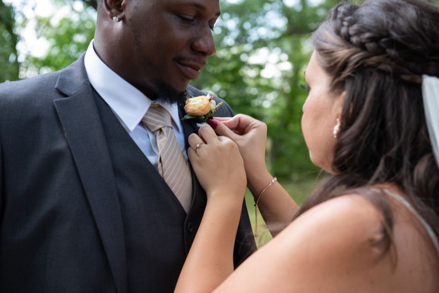 Bride pins boutonniere on groom's lapel during wedding day preparations outdoors.