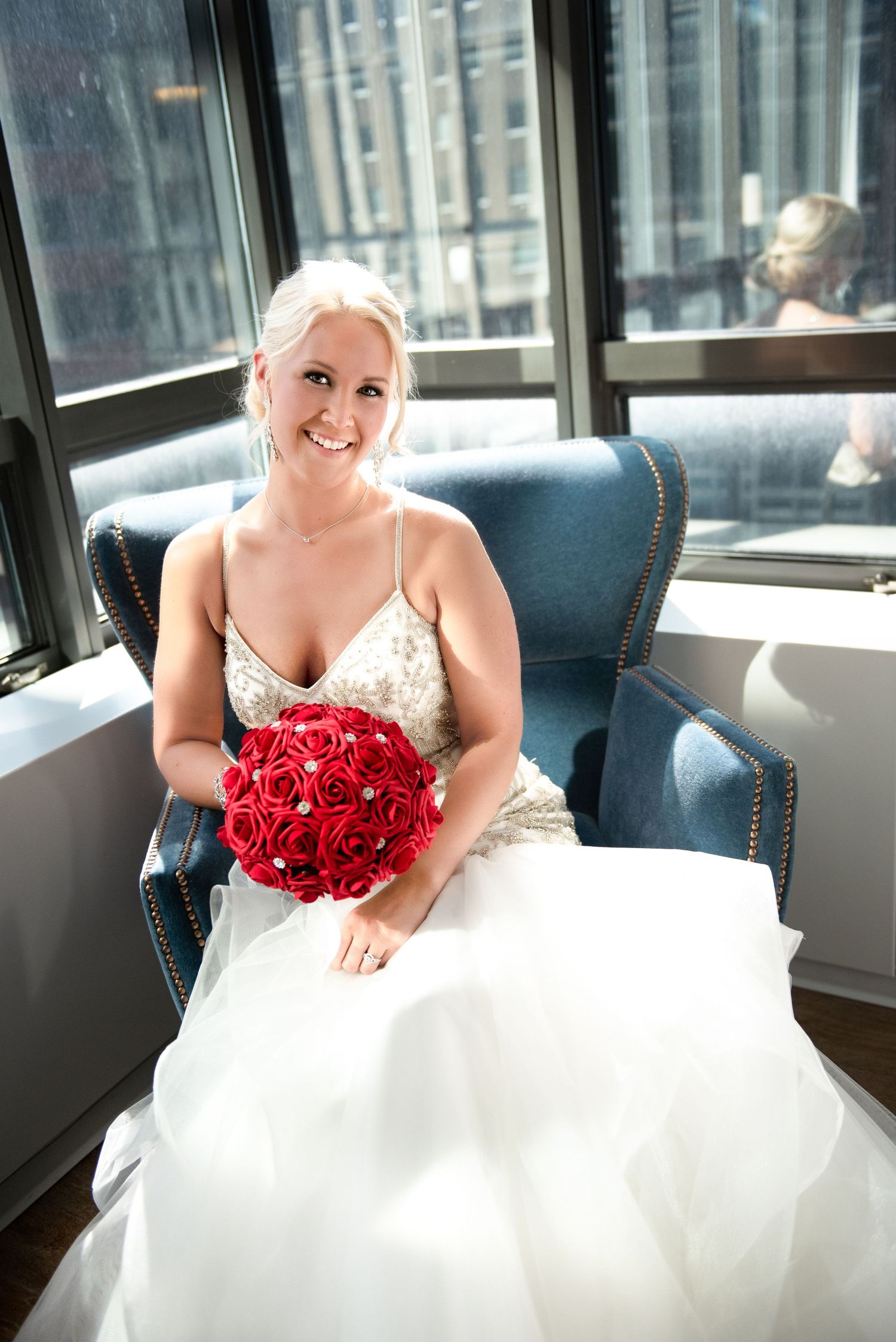 Radiant blonde bride in beaded gown sitting in blue chair holding red rose bouquet near large windows.