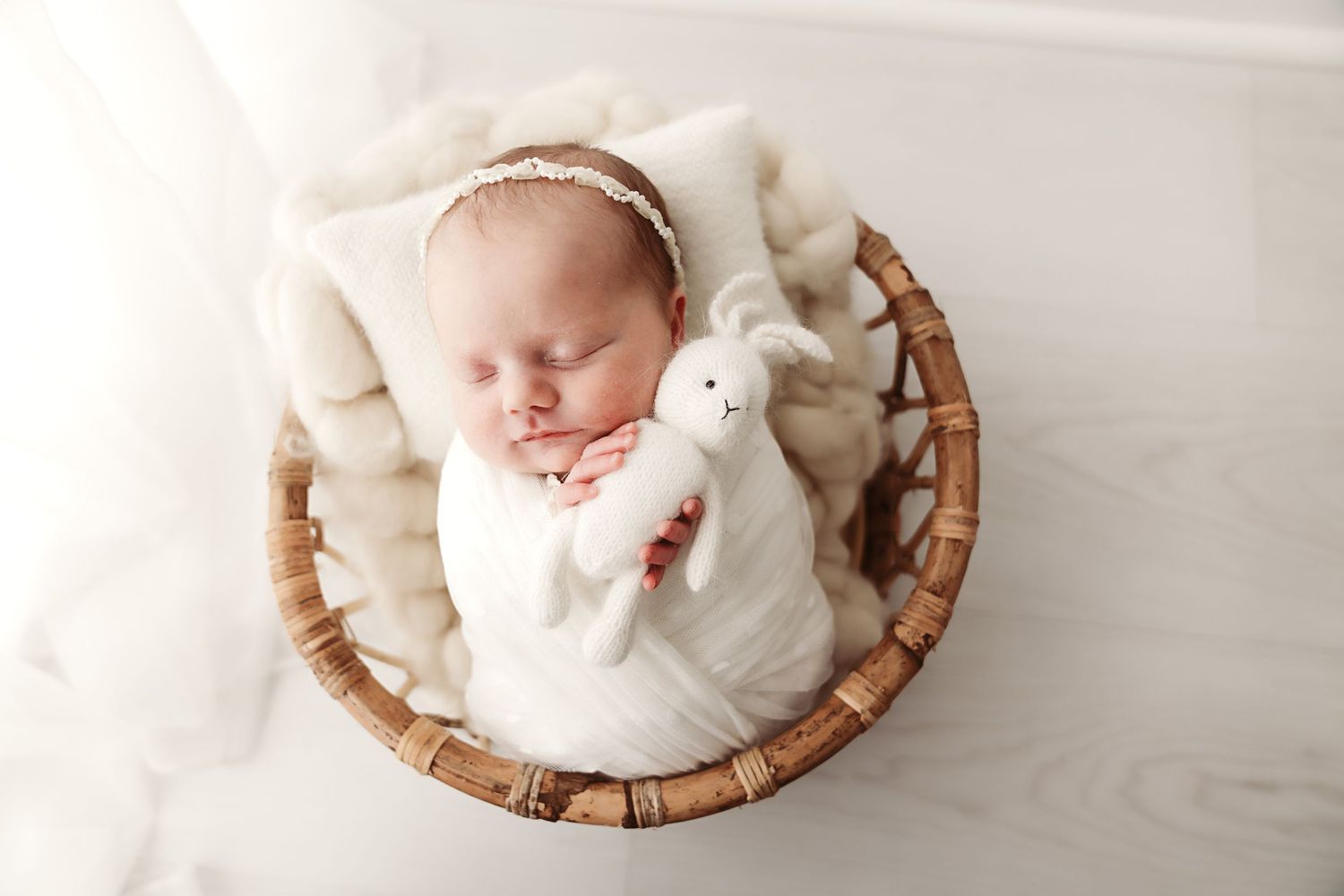 Sleeping newborn baby wrapped in white swaddle holding bunny toy in wicker basket with soft blanket.