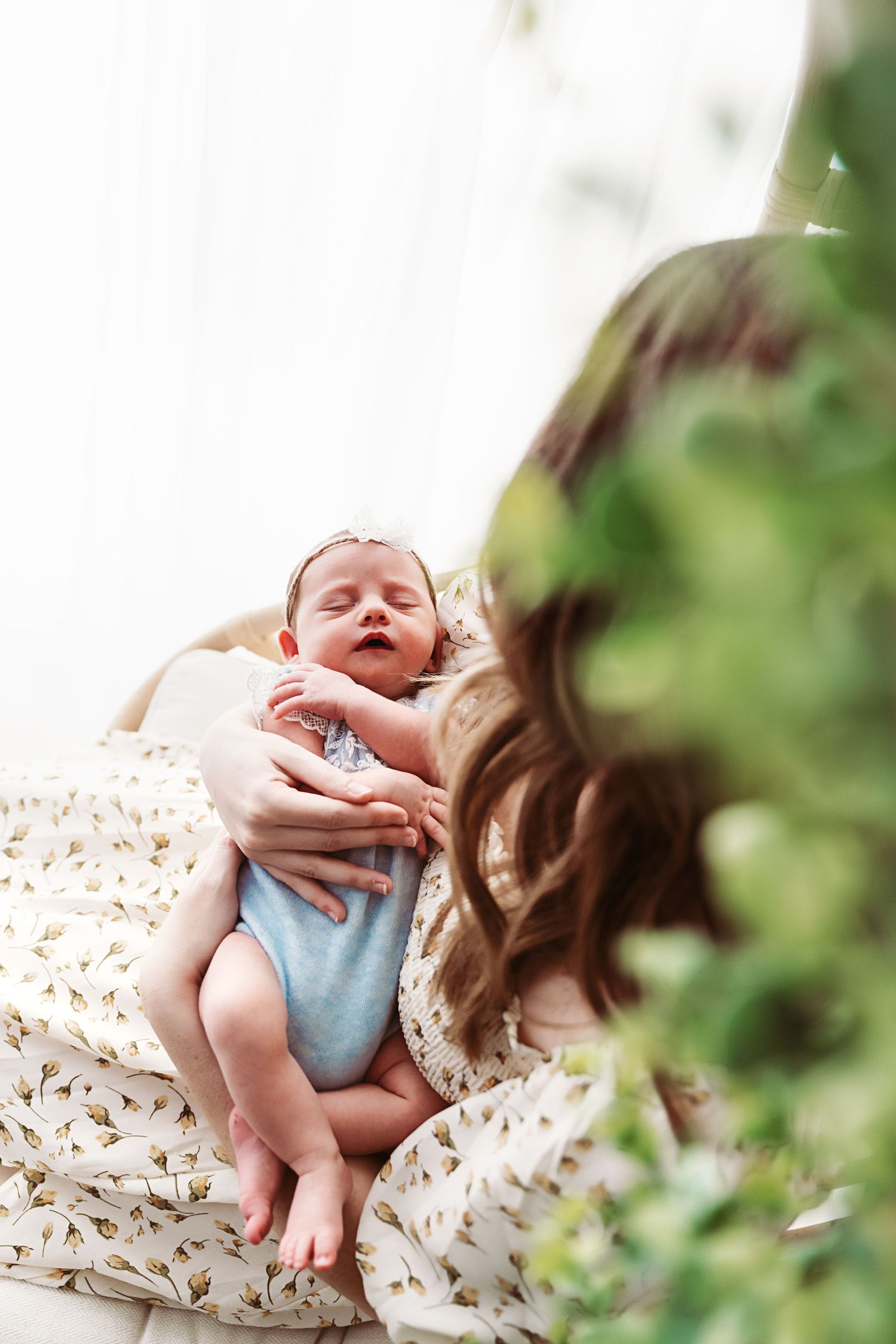 Mother in floral dress cradling her sleeping newborn baby surrounded by green foliage indoors.
