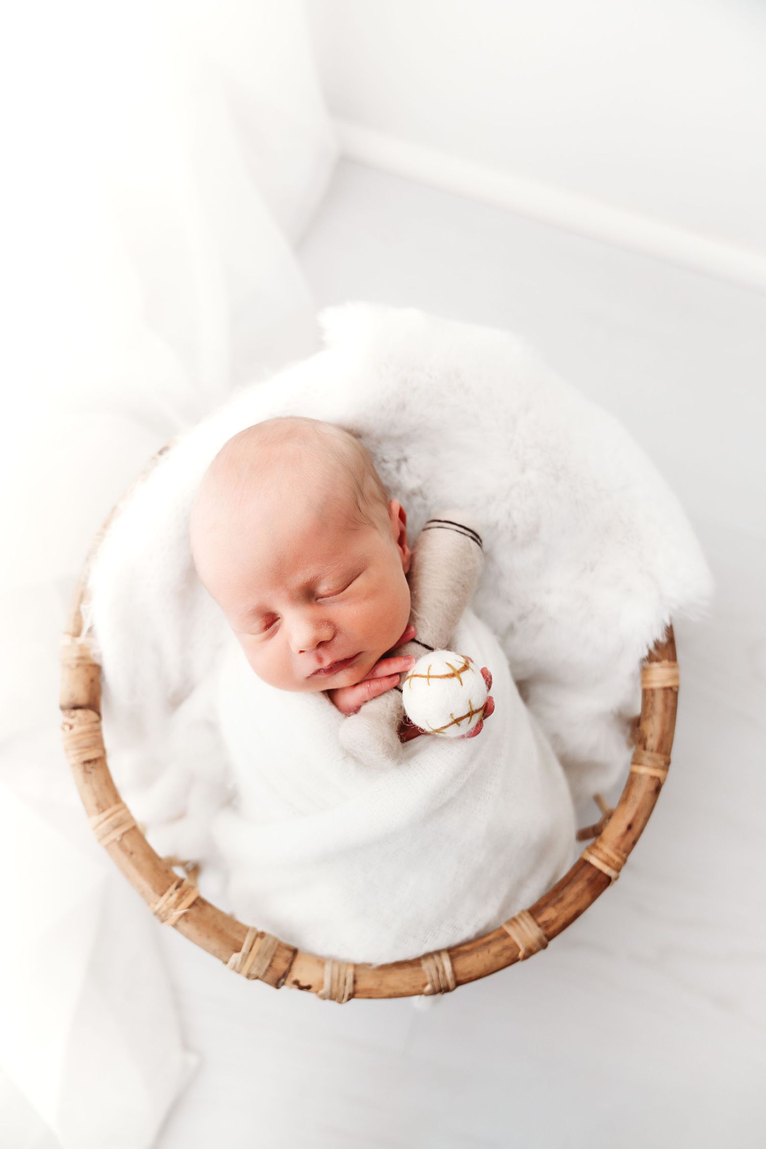 Sleeping newborn baby wrapped in white blanket holding small toy in wicker basket.