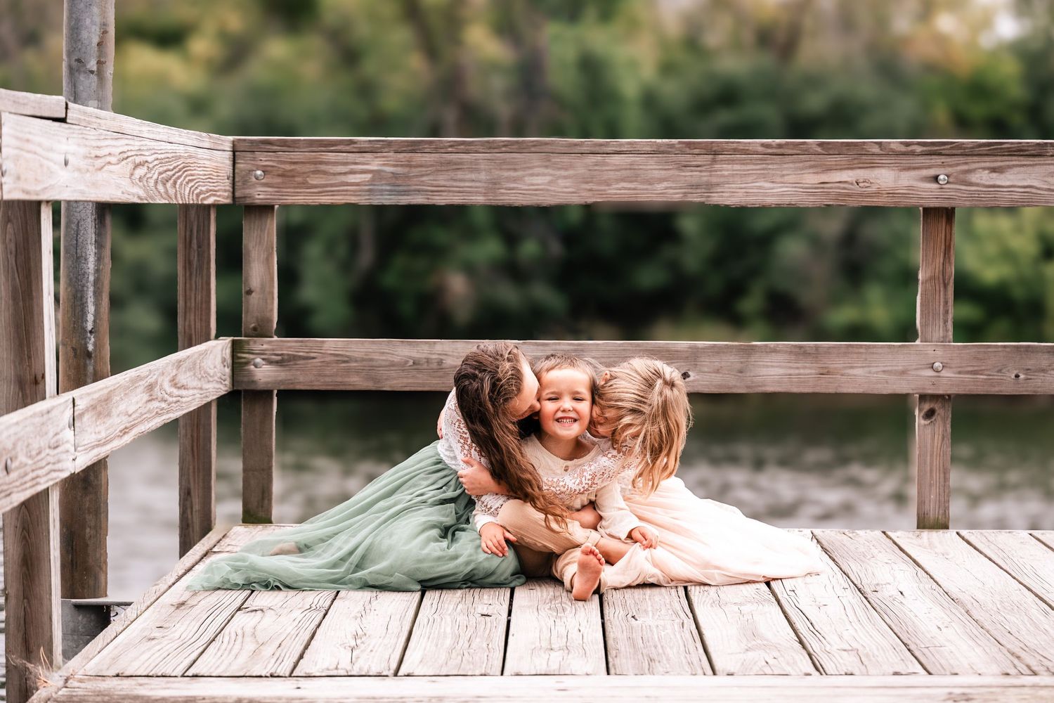 Three children hugging and laughing together on a rustic wooden dock outdoors.