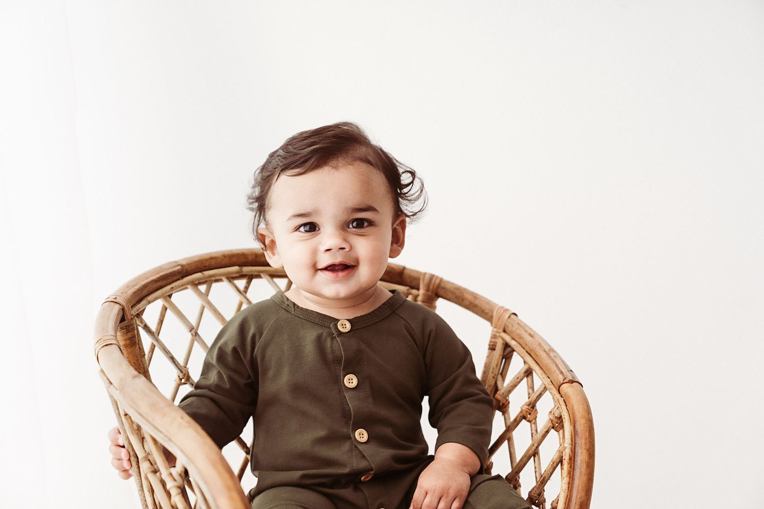 Smiling baby sitting in a round wicker chair wearing an olive green outfit.