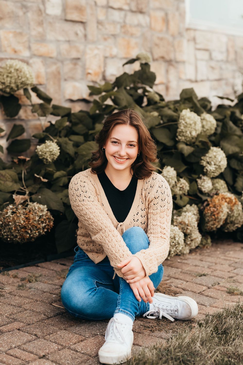 Young woman smiling, sitting on brick pavers in jeans and beige cardigan, surrounded by hydrangea bushes against stone wall.