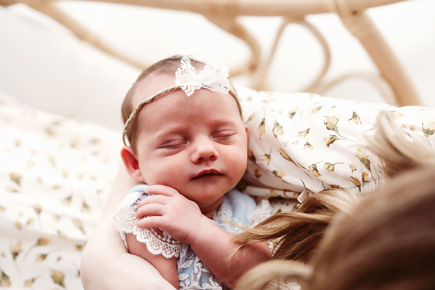 A newborn baby with a floral headband sleeps peacefully in a mother's arms during a professional photo session.