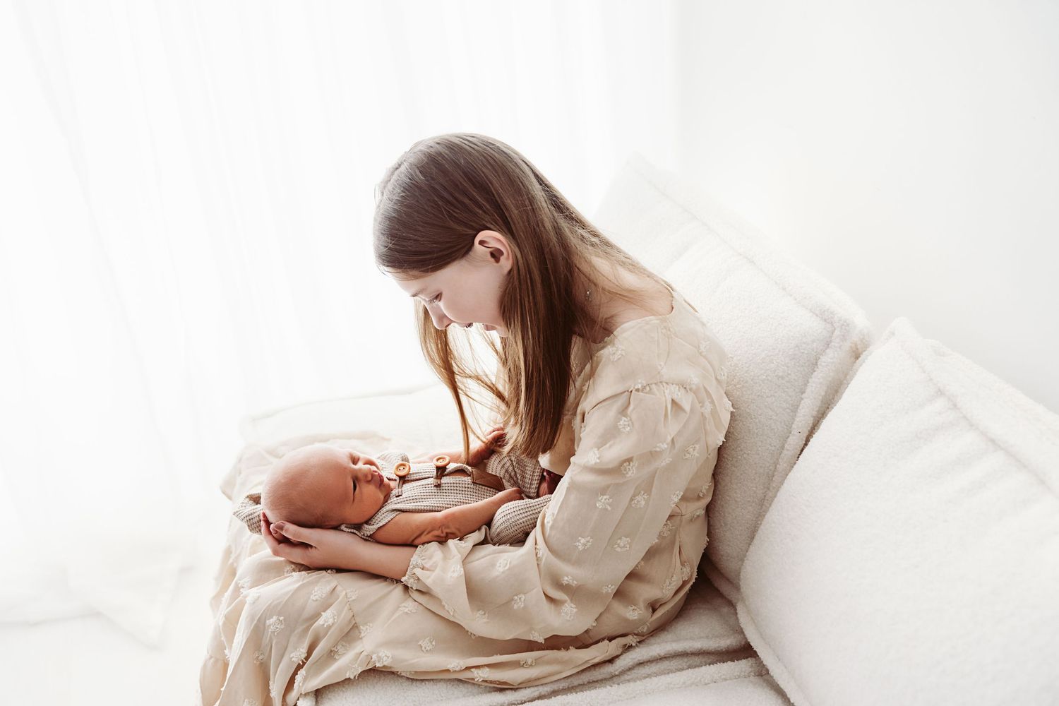 Young girl in beige dress tenderly holds sleeping newborn baby on white sofa.