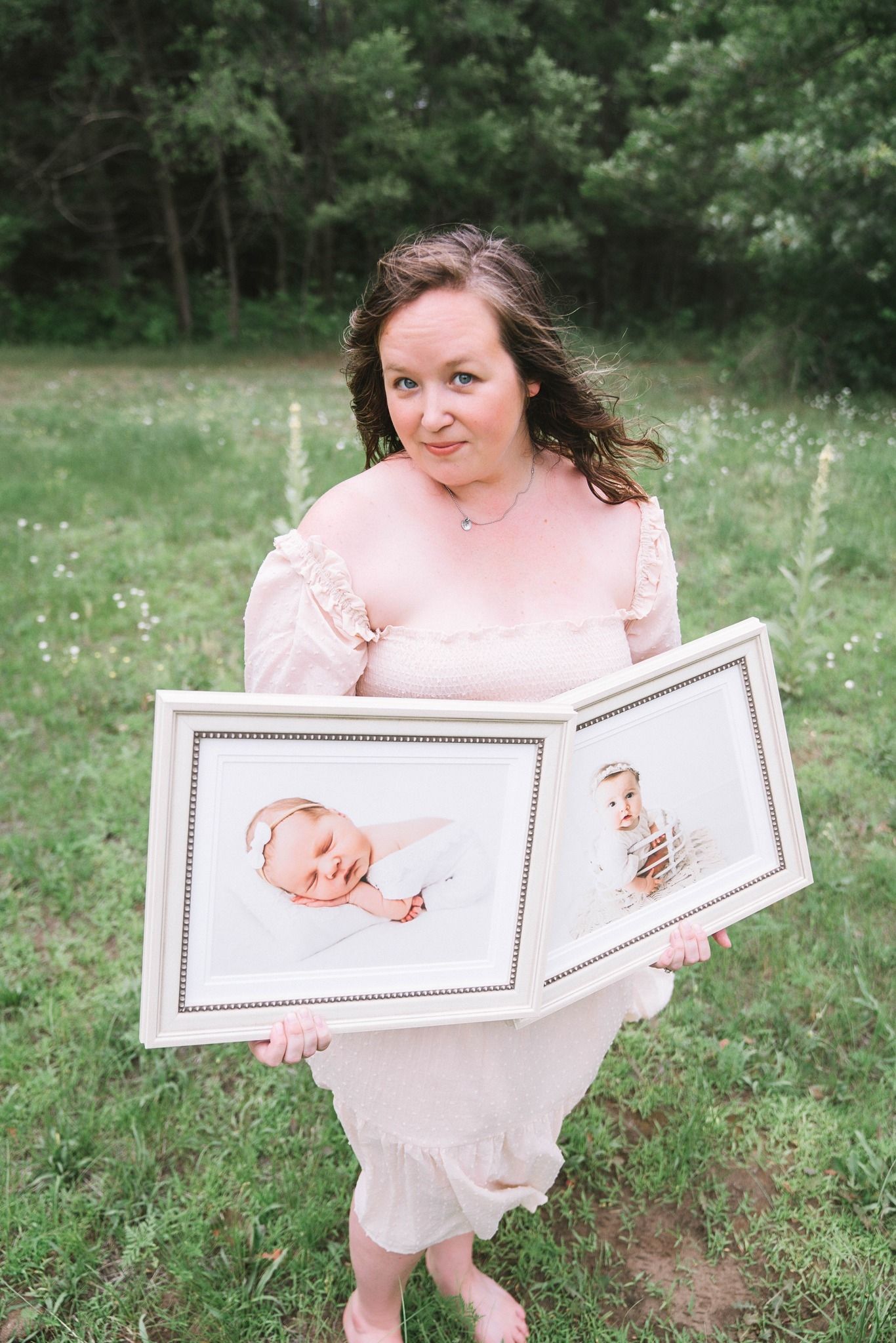 Woman in a pink dress holding an open framed photo album with baby portraits in a green field.