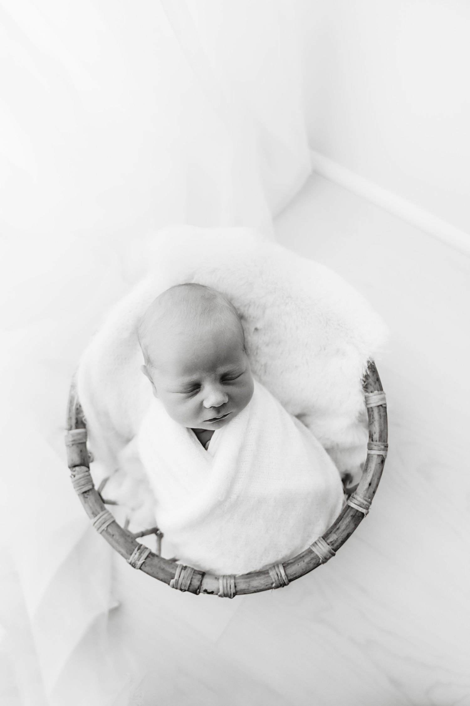 Peaceful newborn baby swaddled in white wrap sleeping in a wicker basket, black and white photo.