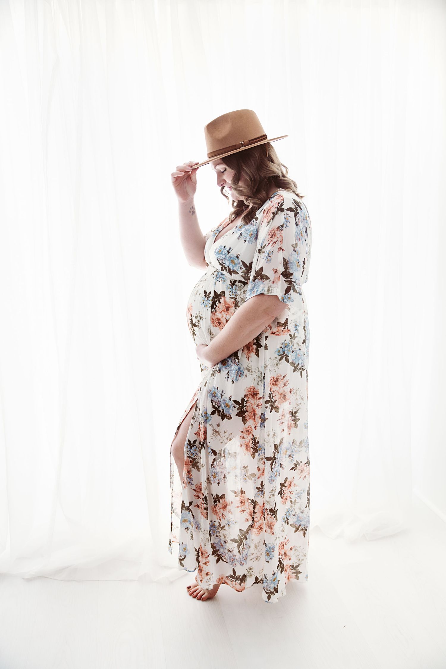 Pregnant woman in floral maxi dress and tan hat posing gracefully in a bright white studio.