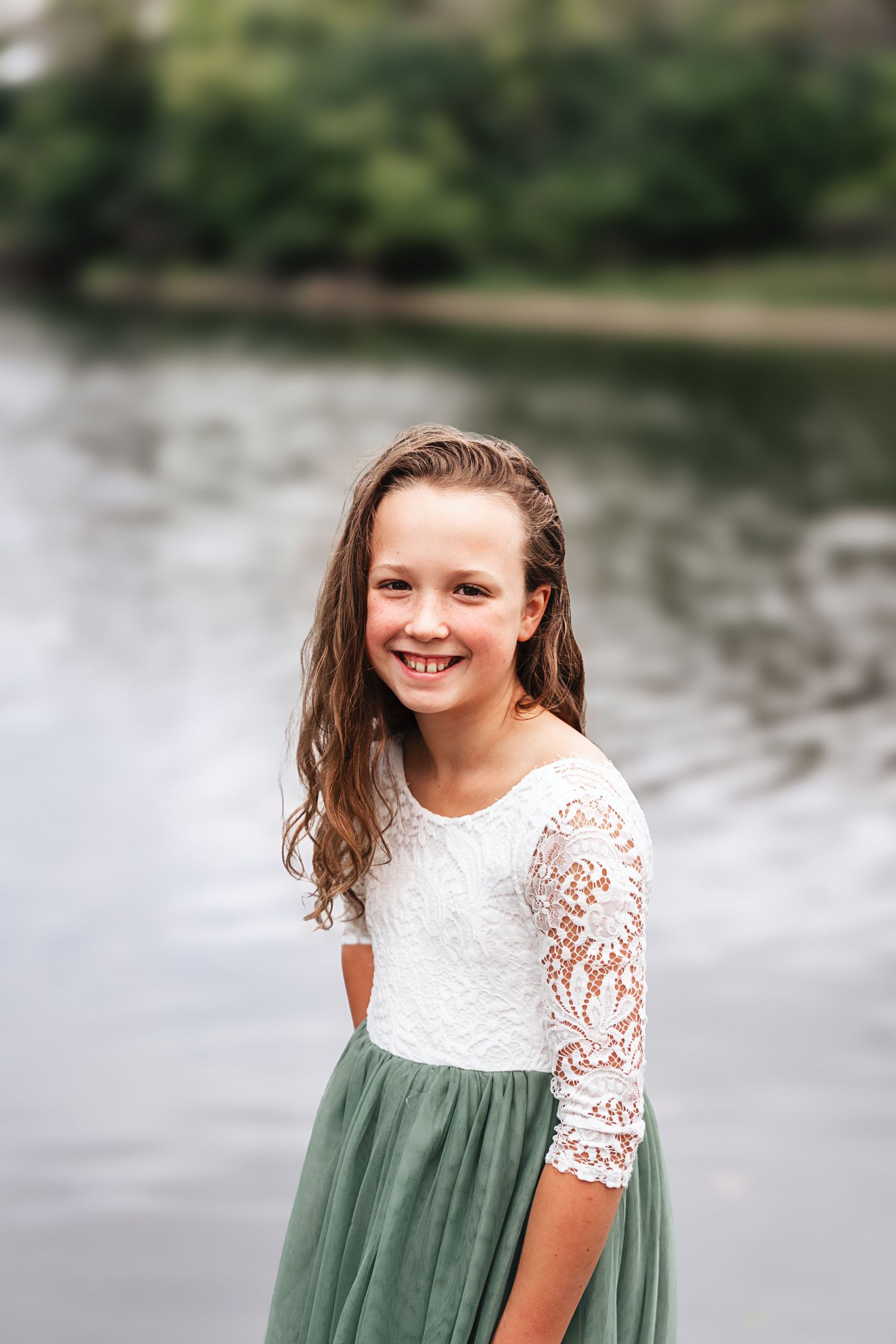 Smiling girl in white lace top and green tulle skirt posing near a river.