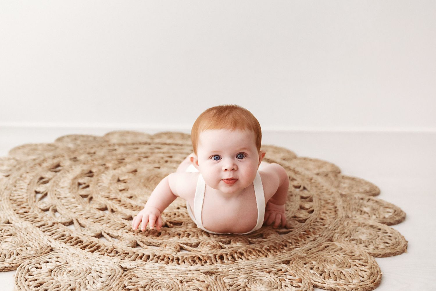 An adorable red-haired baby lies on their tummy on a large woven jute rug.