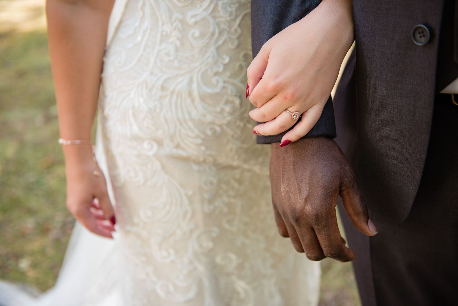 Close-up of interracial couple holding hands, bride wearing gold wedding band outdoors.