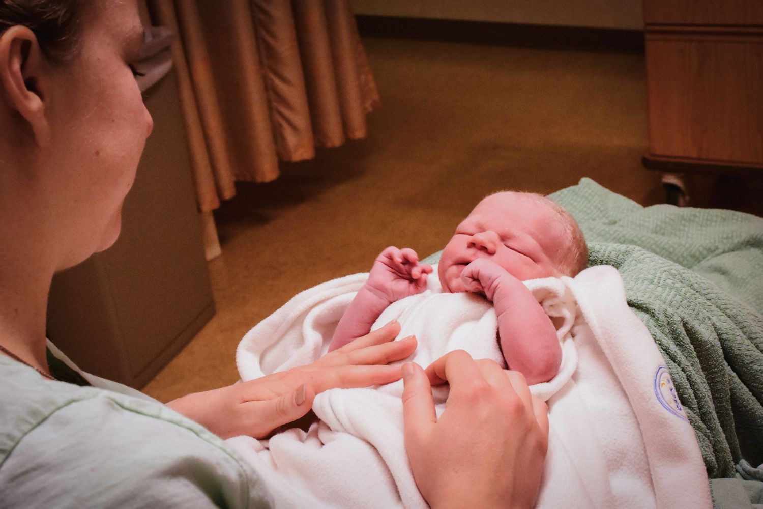 New mother in hospital gown bonding with her crying newborn baby wrapped in white blanket.